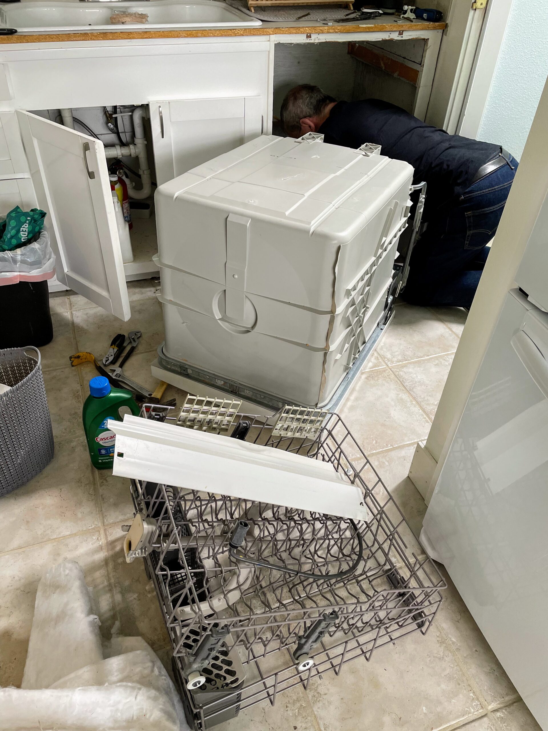 Dishwasher on kitchen floor and man peering into hole under counter where dishwasher used to be