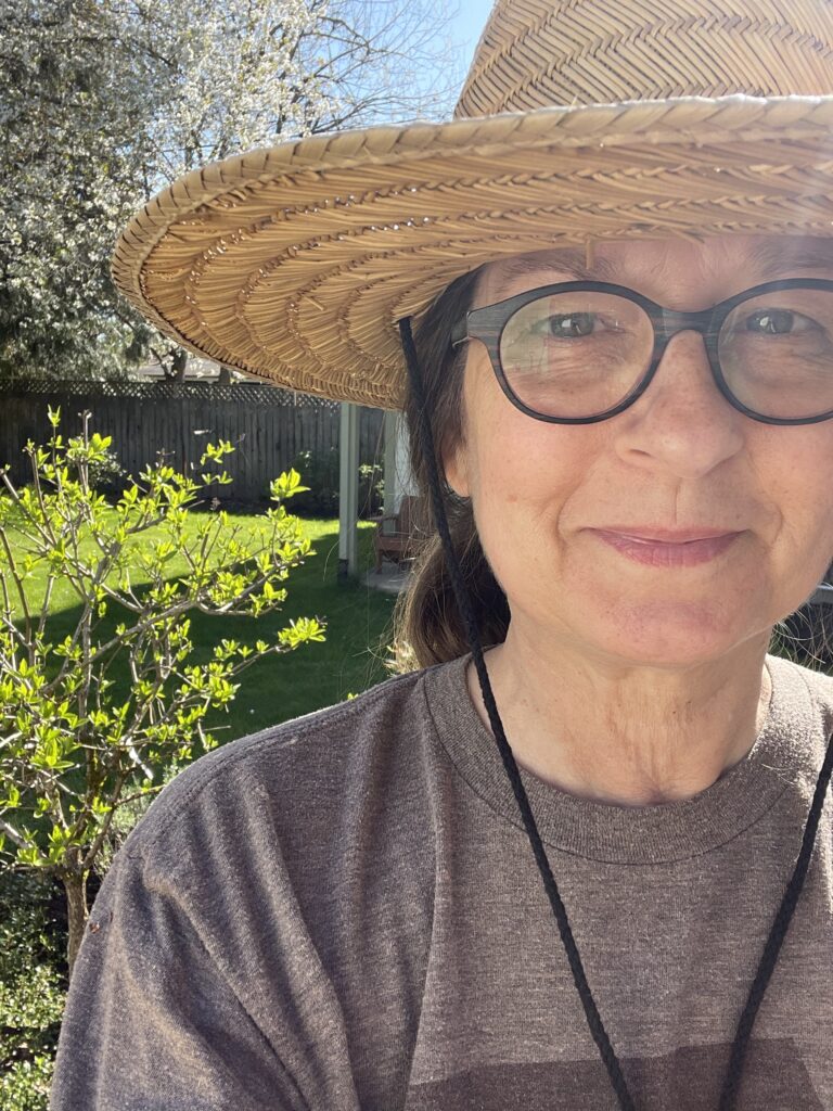 50-something woman in an old t-shirt and gardening hat, smiling, with trees blooming in the background
